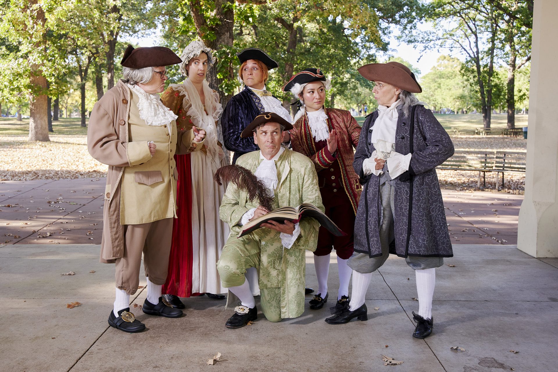 MORTON cast gathered around an actor with a book and quill under a park pavilion
